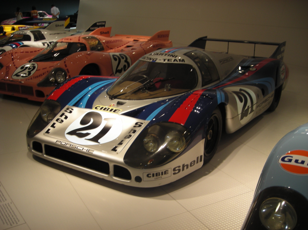 Ein Porsche 917K Rennwagen vom 24-Stunden-Rennen von Le Mans in einem Museum neben anderen Sportwagen ausgestellt, mit Menschen, die zuschauen, und einer Wand im Hintergrund.