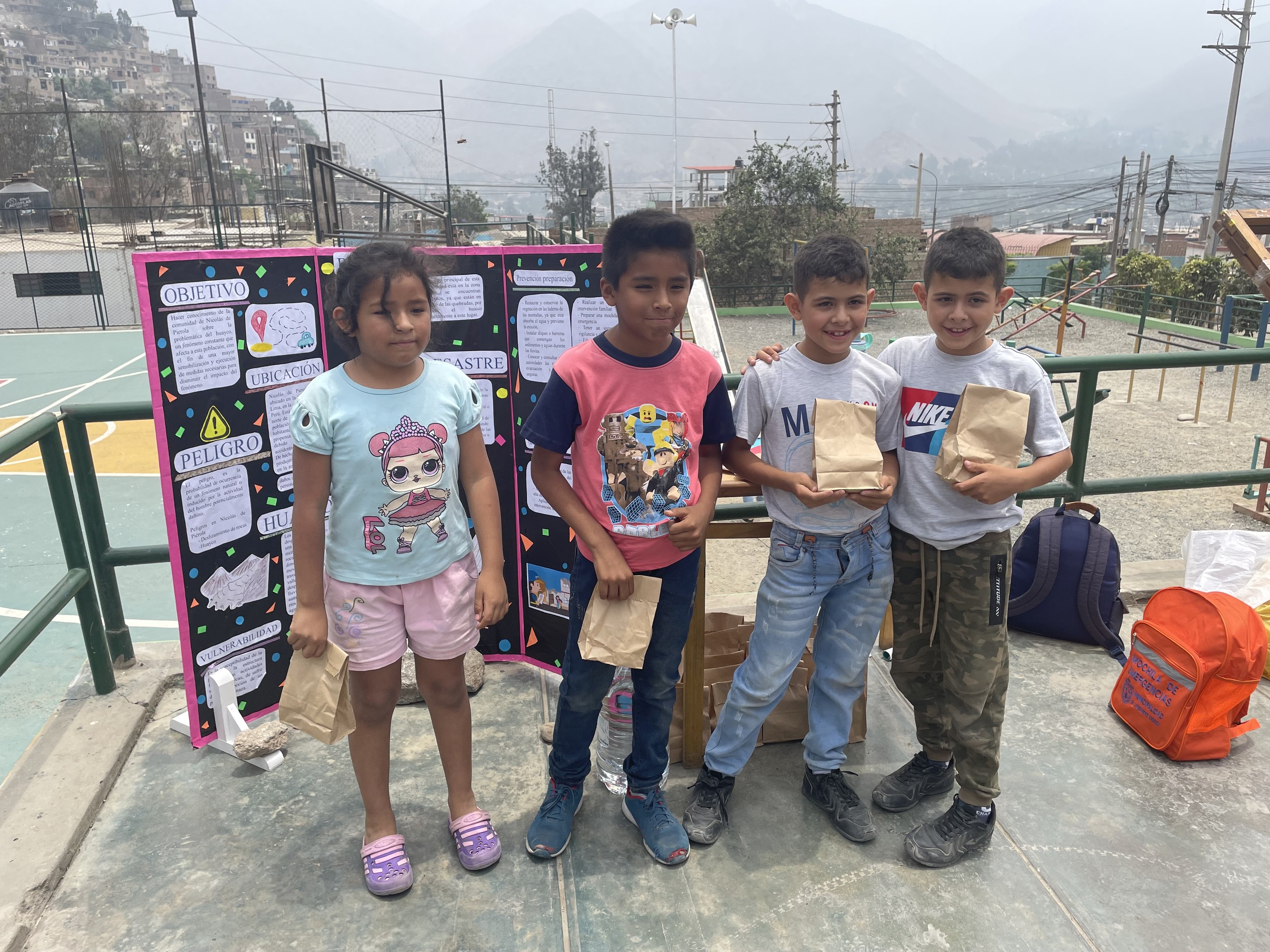 Gruppe von Kindern auf einem Basketballfeld mit Papiertüten in den H├Ąnden, mit Tüten und Gegenst├Ąnden auf der rechten Seite und einer Tafel mit Text auf der linken Seite, vor dem Hintergrund von Z├Ąunung, Masten, Stra├čenlaternen, B├Ąumen, Geb├Ąuden, H├Ęllen und Himmel.