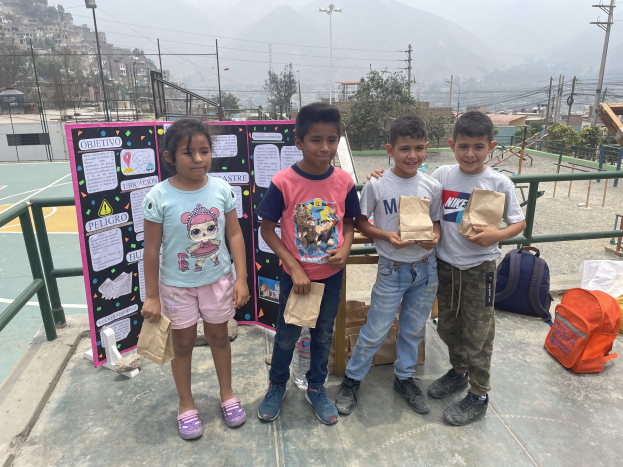 Gruppe von Kindern auf einem Basketballfeld mit Papiertüten in den H├Ąnden, mit Tüten und Gegenst├Ąnden auf der rechten Seite und einer Tafel mit Text auf der linken Seite, vor dem Hintergrund von Z├Ąunung, Masten, Stra├čenlaternen, B├Ąumen, Geb├Ąuden, H├Ęllen und Himmel.