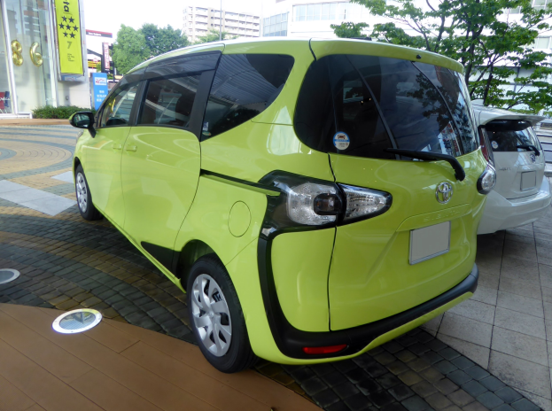 Kleiner grüner Elektroauto vor einem Gebäude mit Bäumen, Pflanzen und Schildern im Hintergrund unter einem sichtbaren Himmel geparkt.