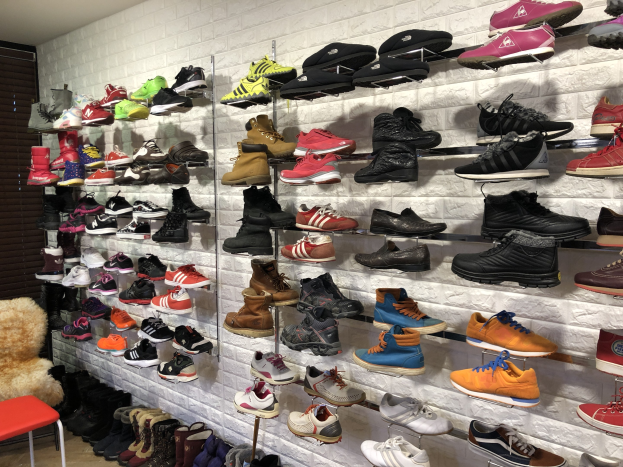 Ein Schuhgeschäft mit zahlreichen Schuhen auf den Gestellen, einem Stuhl mit einem Teddybär auf der linken Seite und einer Wand im Hintergrund.