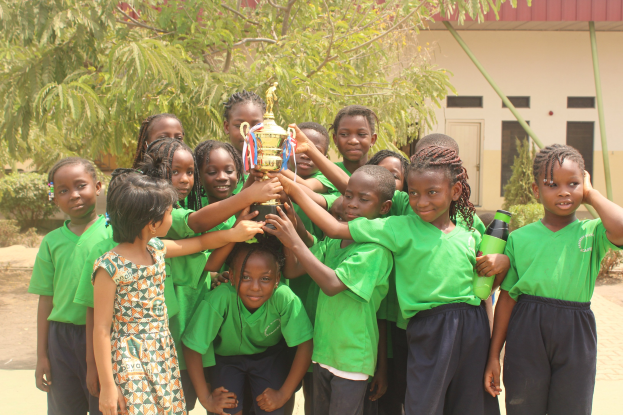 Gruppe von Kindern mit Pokal in Schulhof mit Bäumen und Gebäude im Hintergrund