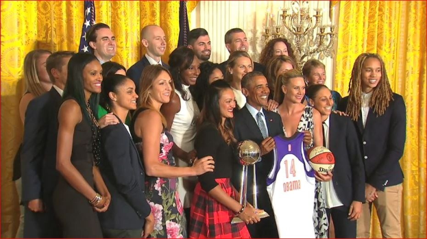 Präsident Obama und First Lady Michelle Obama posieren mit dem Frauen-Basketball-Team im Oval Office des Weißen Hauses, halten einen Basketball, eine Trophäe und lächeln in der Nähe einer Flagge, von Vorhängen und einer Kerzenleuchter.