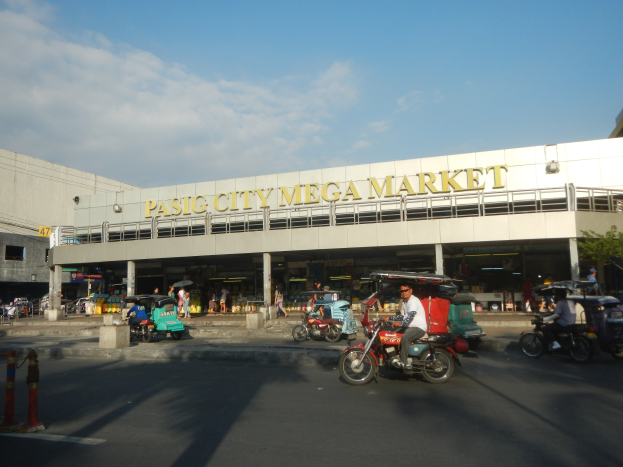 Eine belebte Straßenszene am Pasig City Mega Market mit Fahrzeugen und Fußgängern, einem Gebäude mit Text im Hintergrund und einer bewölkten Himmel.