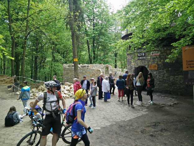 People walking down a wooded path, some carrying bicycles and wearing helmets, with a stone wall and a sign indicating the entrance to the Szyb Stylweste Mountain Bike Trail in the background.