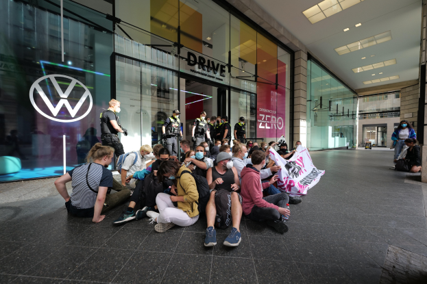 Menschen sitzen auf dem Boden vor einer Volkswagen-Filiale und halten ein Transparent.