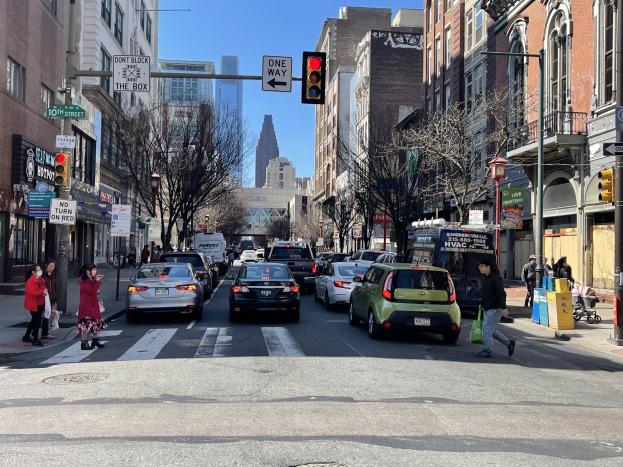 Eine belebte Stadtstraße mit Fahrzeugen, Fußgängern, Bäumen, Gebäuden, Schildern, Laternenpfählen, einer Ampel und einem geparkten Van unter einem sichtbaren Himmel.