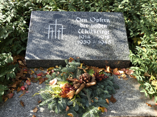 Grabstein mit einem Kränzchen in einem Friedhof, mit Inschrift für Wilhelm Wilhelm umgeben von Pflanzen und Blättern.