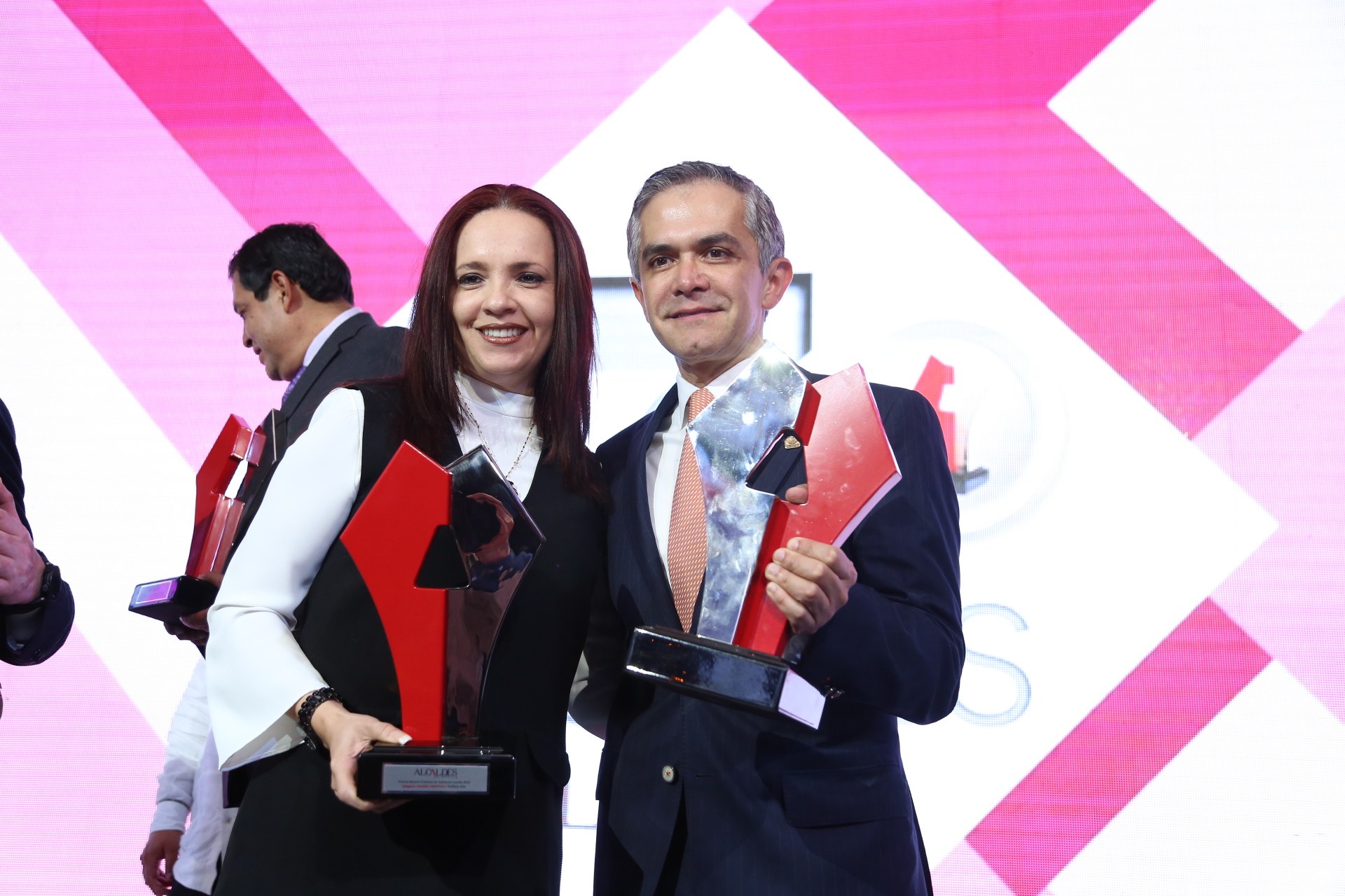 Zwei Personen auf einer Bühne mit Trophäen, die lächeln, und ein Bildschirm im Hintergrund.