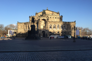 Großer beige Berliner Dom mit einem prominenten Dom und einer Statue davor, umgeben von Straßenlaternen, Fahrzeugen, Fußgängern, Zelten, Bäumen und einem bewölkten Himmel.