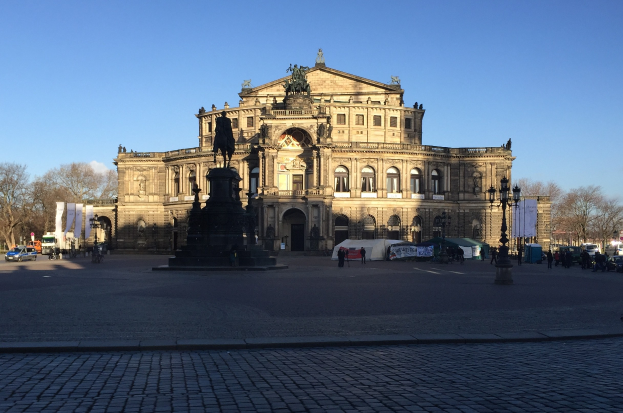 Großer beige Berliner Dom mit einem prominenten Dom und einer Statue davor, umgeben von Straßenlaternen, Fahrzeugen, Fußgängern, Zelten, Bäumen und einem bewölkten Himmel.