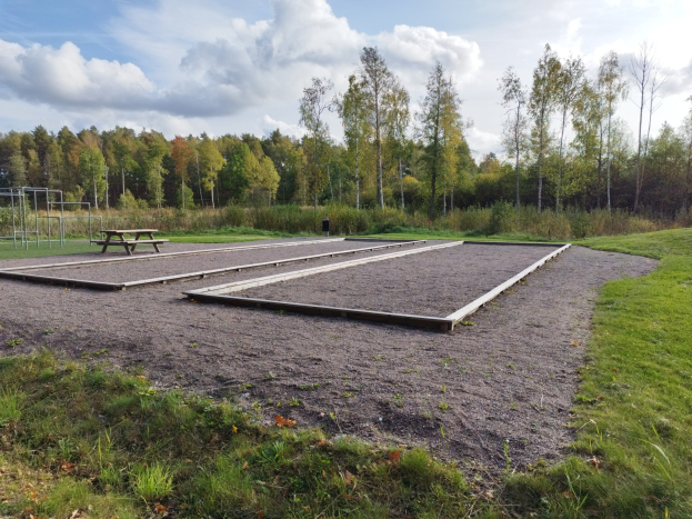 Eine grüne Wiese mit einem Picknicktisch, umgeben von Bäumen unter einem klaren blauen Himmel, mit Spielplatzgeräten wie Stangen und einer Bank auf der linken Seite.