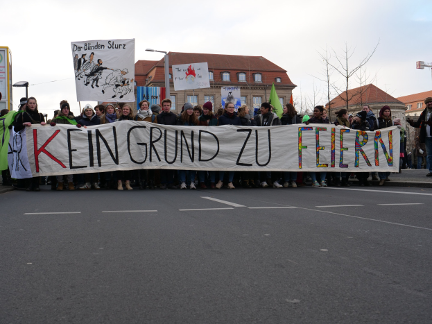 Demonstranten mit einem Banner mit der Aufschrift 'Kein Grund zur Feier' gegen deutsche Sparmaßnahmen, mit Gebäuden, Bäumen und einem klaren Himmel im Hintergrund.