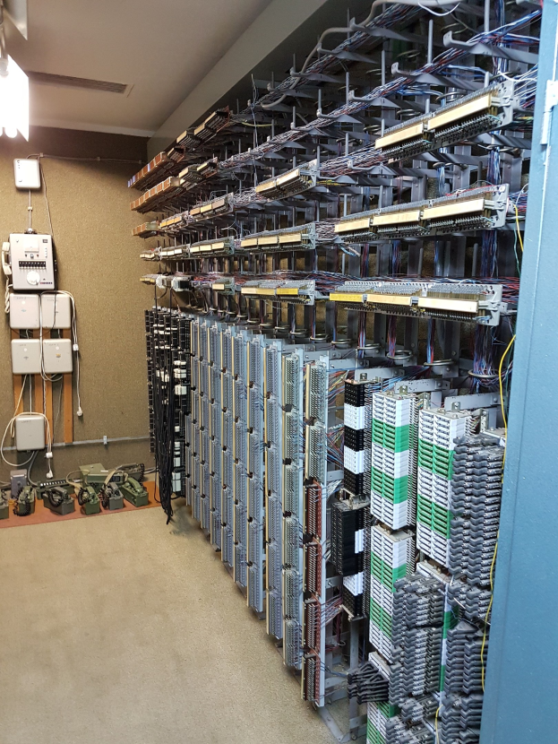 Großer Raum mit zahlreichen Kabeln und elektronischen Geräten auf dem Boden, darunter Server und Kabel, was auf ein Rechenzentrum mit einer sichtbaren Wand auf der linken Seite hinweist.