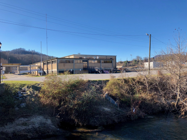 Grüne Wiese mit einem Fluss, Bäumen, Pflanzen, Gras, Steinen, Straßenlaternen, Strommasten, Stromkabeln, Hügeln, einem klaren blauen Himmel und einem Gebäude an der Stelle einer geplanten Wasseraufbereitungsanlage.