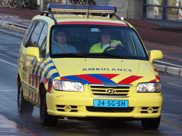 Ein gelber Krankenwagen mit zwei Insassen fährt eine Straße entlang, mit parkenden Fahrrädern auf dem linken Gehweg und einem Gebäude mit Glasfenstern im Hintergrund.