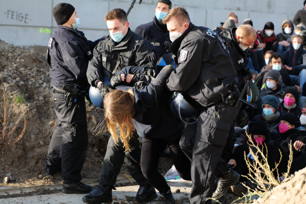Gruppe maskierter Personen in Kapuzenpullis, einige bewaffnet, die eine Frau umgeben, die von der Polizei begleitet wird, mit Pflanzen, Steinen und einer Wand mit Protesttext im Hintergrund.