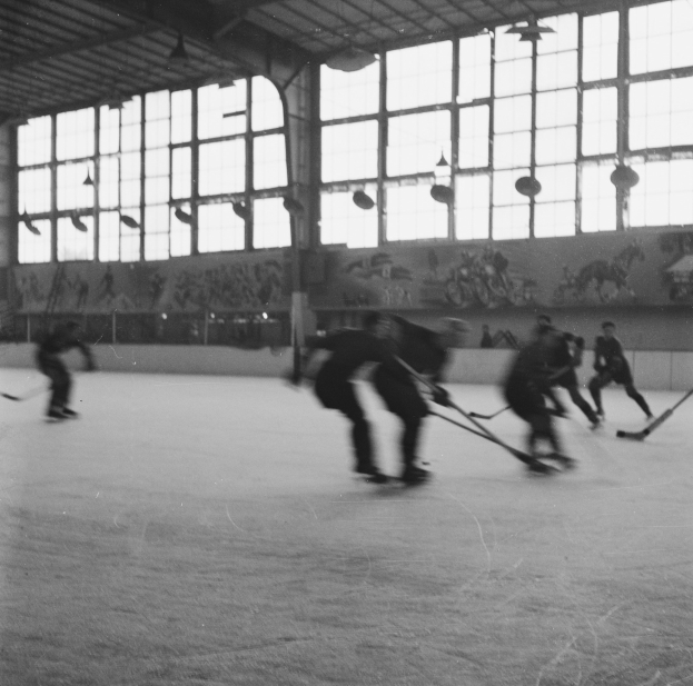 Schwarzes und weißes Foto von Menschen, die Hockey auf einem Eisplatz spielen, mit Gemälden und Fenstern an der Wand dahinter.