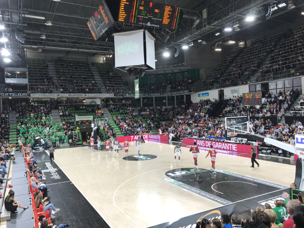 Basketballspiel in einer großen Halle mit Zuschauern, Texttafeln mit "Asvel vs Galatasaray" und Decke mit Lampen und Bildschirm.