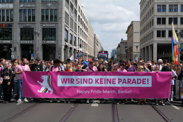 Eine Gruppe von Menschen, die eine rosafarbene Fahne mit der Aufschrift "Happy Pride March" schwenken, geht eine Stra√e in Berlin, Deutschland, entlang, mit Gebäuden, Laternenmästen und Verkehrszeichen an der Stra√e und einem bewölktem Himmel im Hintergrund.