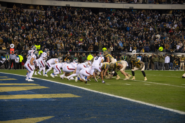Ein Fuballspiel im Gange mit Spielern in Sportkleidung und Helmen auf dem Feld, Zuschauern, Gelöndern und Tafeln mit Text im Hintergrund.