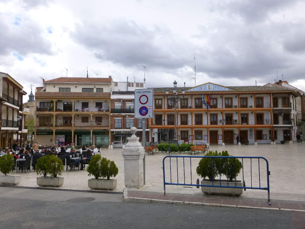Ein belebter Stadtplatz mit Menschen auf St"{u}hlen und Stehenden, umgeben von Topfpflanzen, Metallabsperrungen, Stra{\ss}enmasten mit Flaggen, ein Schild, Geb"{a}ude mit Fenstern und einem bew"{o}lktem Himmel.