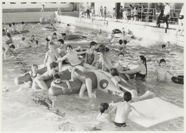 Schwarzes und weißes Bild von Kindern, die in einem Swimmingpool spielen, einige auf aufblasbaren Gegenständen und andere stehend, mit einer Wand und einem Geländer im Hintergrund.