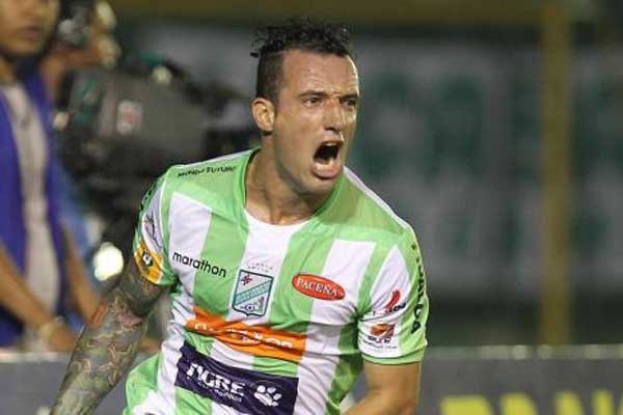 Ein Fußballspieler in grüner und weißer Uniform mit Tattoos auf dem Arm feiert nach einem Tor, mit unscharfem Hintergrund und einer Tafel.