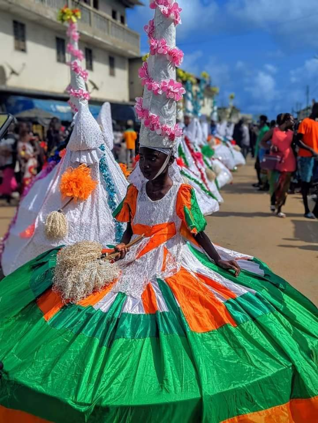 Eine Gruppe von Menschen in bunten Kostümen geht eine Straße entlang, mit einem bewölkten Himmel und einem Gebäude im Hintergrund, wahrscheinlich bei einem festlichen Ereignis.
