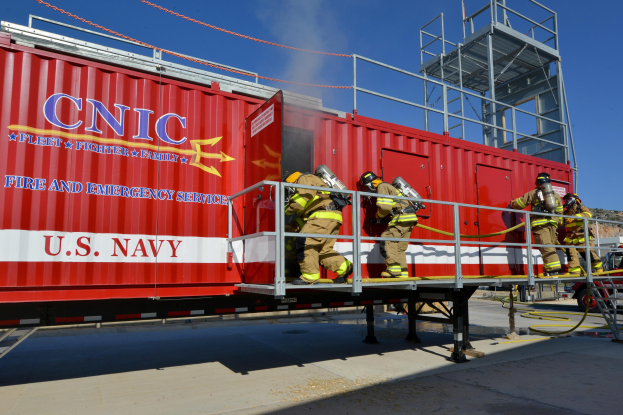 Feuerwehrleute mit Helmen stehen auf einem roten Seecontainer mit Text, halten Rohre in der Nähe von Geländern, Treppen und einem Fahrzeug, mit Bäumen und bewölktem Himmel im Hintergrund.