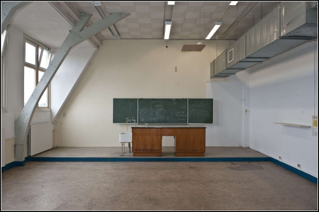 Ein leeres Klassenzimmer mit einer Tafel, einem Tisch, einem Waschbecken mit Wasserhahn, einem Hocker, einem Fenster und einer Deckenbeleuchtung.