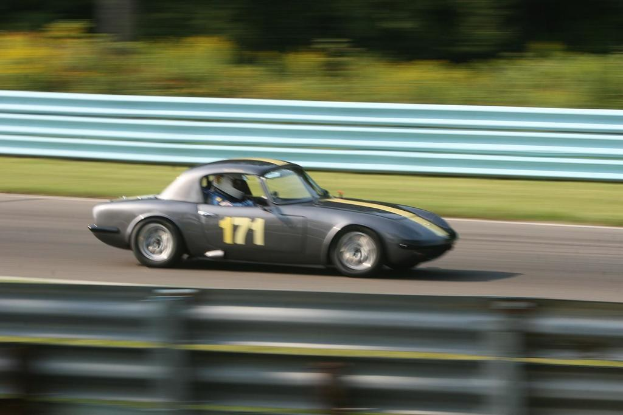 Ein Auto mit einem Helm tragenden Fahrer fährt eine Straße entlang, die von Barrieren gesäumt ist und Bäume im Hintergrund hat.