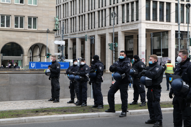 Eine Gruppe von Polizisten in schwarzen Uniformen und Masken steht vor einem Gebäude mit Glasfenstern und Säulen in Berlin, Deutschland, einige halten Helme, mit Laternenmasten, Verkehrsampeln, Schildern mit Text, einer Statue und anderen Menschen im Hintergrund.