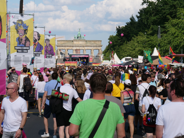 Eine große Menschenmenge geht eine Straße vor dem Brandenburger Tor in Berlin, Deutschland entlang, viele tragen Mützen und Taschen, Banner links, Bäume, Zelte, Luftballons und ein Tor im Hintergrund bei bewölktem Himmel.