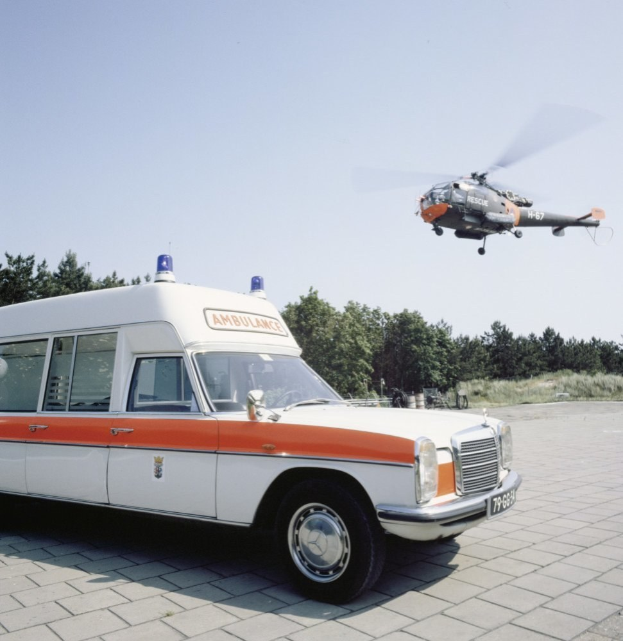 Rettungswagen vor einem Hubschrauber mit Bäumen und einem klaren blauen Himmel im Hintergrund.