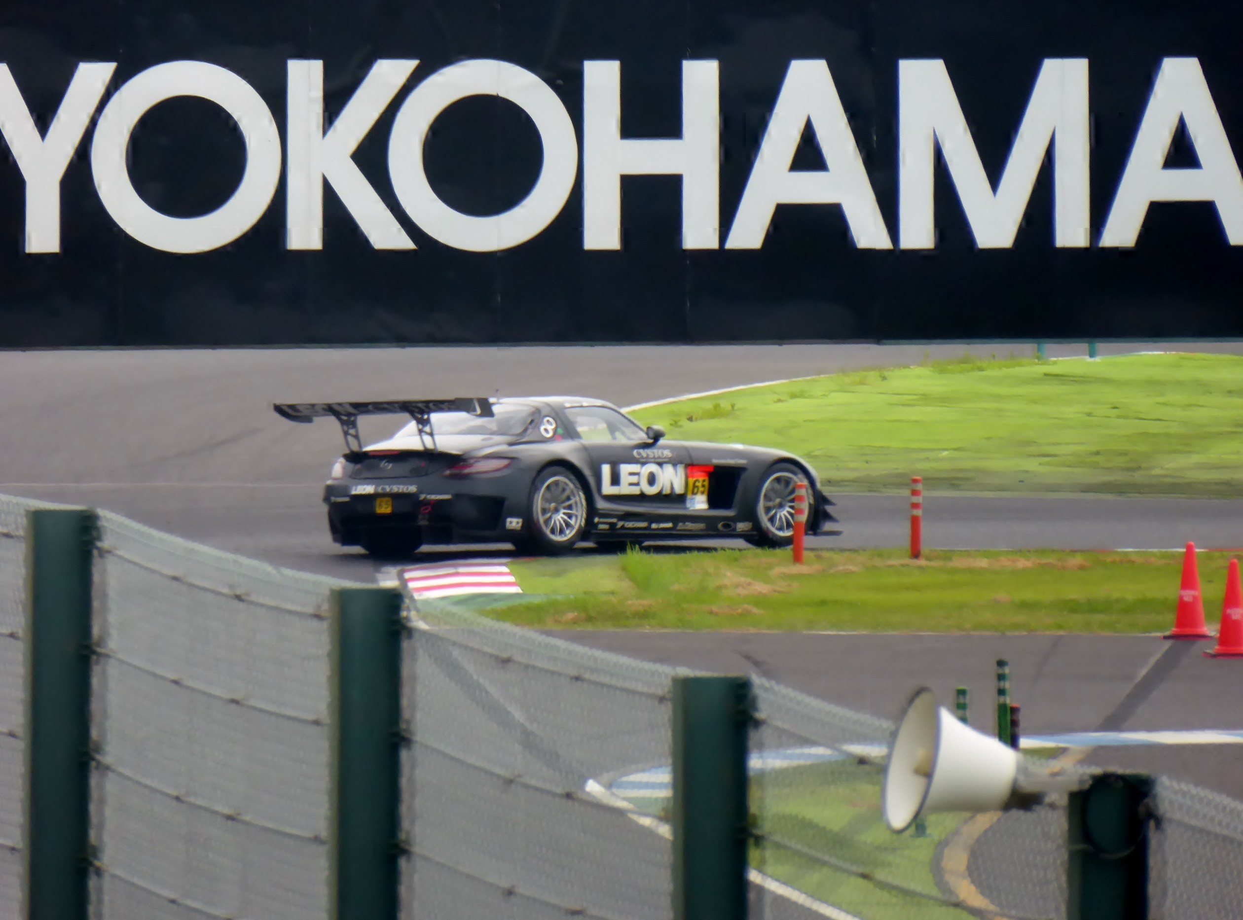 Schwarzer Sportwagen auf einer Rennstrecke mit Verkehrsleitkegeln umgeben, mit einem "Yokohama"-Schild im Hintergrund, Gras, einem Zaun und einem Lautsprecher im Vordergrund.
