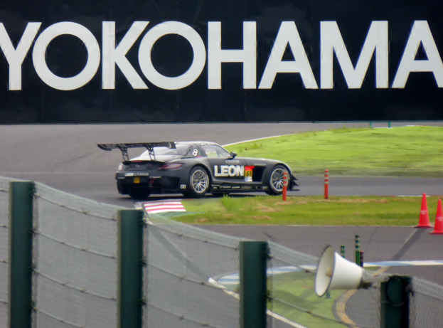 Schwarzer Sportwagen auf einer Rennstrecke mit Verkehrsleitkegeln umgeben, mit einem "Yokohama"-Schild im Hintergrund, Gras, einem Zaun und einem Lautsprecher im Vordergrund.