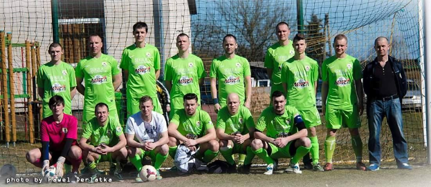Eine Gruppe von Männern in grünen T-Shirts steht auf einem Fußballfeld, einige in Hockstellung, mit einem Ball und einem Torpfosten; der Hintergrund zeigt Bäume, Gebäude und Fahrzeuge.