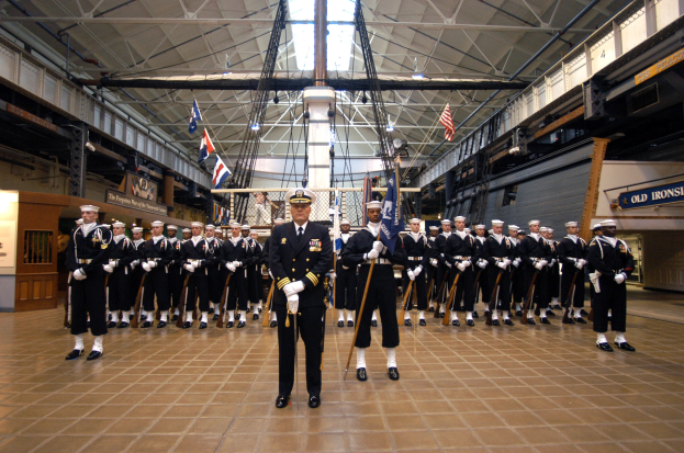 Gruppe uniformierter Männer in einem großen Raum mit Gewehren, Fahnen, Schildern und Deckenleuchten und Stangen.