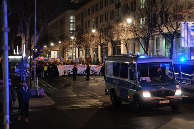 Eine Gruppe von Menschen, die nachts eine Straße entlanggehen, mit einem Polizeiwagen auf der Seite und einem Banner im Hintergrund.