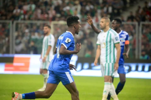 Ein Fußballspieler in einem blauen und weißen Trikot feiert nach einem Torerfolg, umgeben von einer Gruppe von Menschen in weißen und grünen Trikots, mit einem weißen und blauen Brett und Geländer im unscharfen Hintergrund.