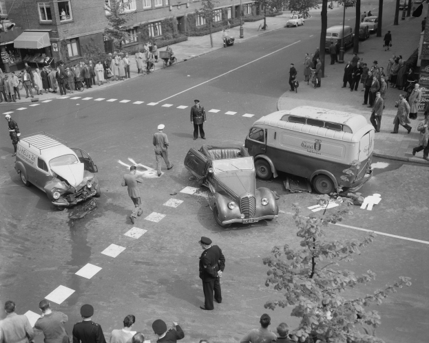 Schwarz-weiß-Szene eines Autounfalls an einer Straßenkreuzung mit einem Lieferwagen, anderen Fahrzeugen und Menschen in der Nähe, mit Gebäuden, Bäumen, Pfählen und Texttafeln im Hintergrund.
