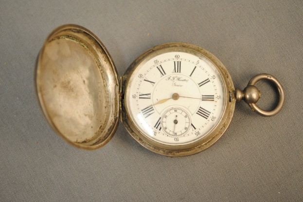 Eine silberne Taschenuhr mit einem weißen Zifferblatt mit römischen Zahlen, geschlossenem Deckel, auf einer grauen Oberfläche.