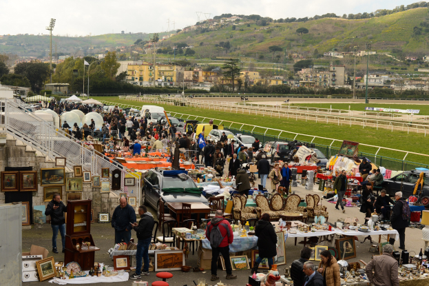 Große Gruppe von Menschen auf einem Flohmarkt mit Tischen, auf denen Gegenstände wie Foto Rahmen und Stühle ausgelegt sind, Fahrzeuge in der Nähe geparkt, Geländer, Treppen, Bäume, Gebäude, Laternenpfähle, Hügel und ein bewölkter Himmel im Hintergrund.