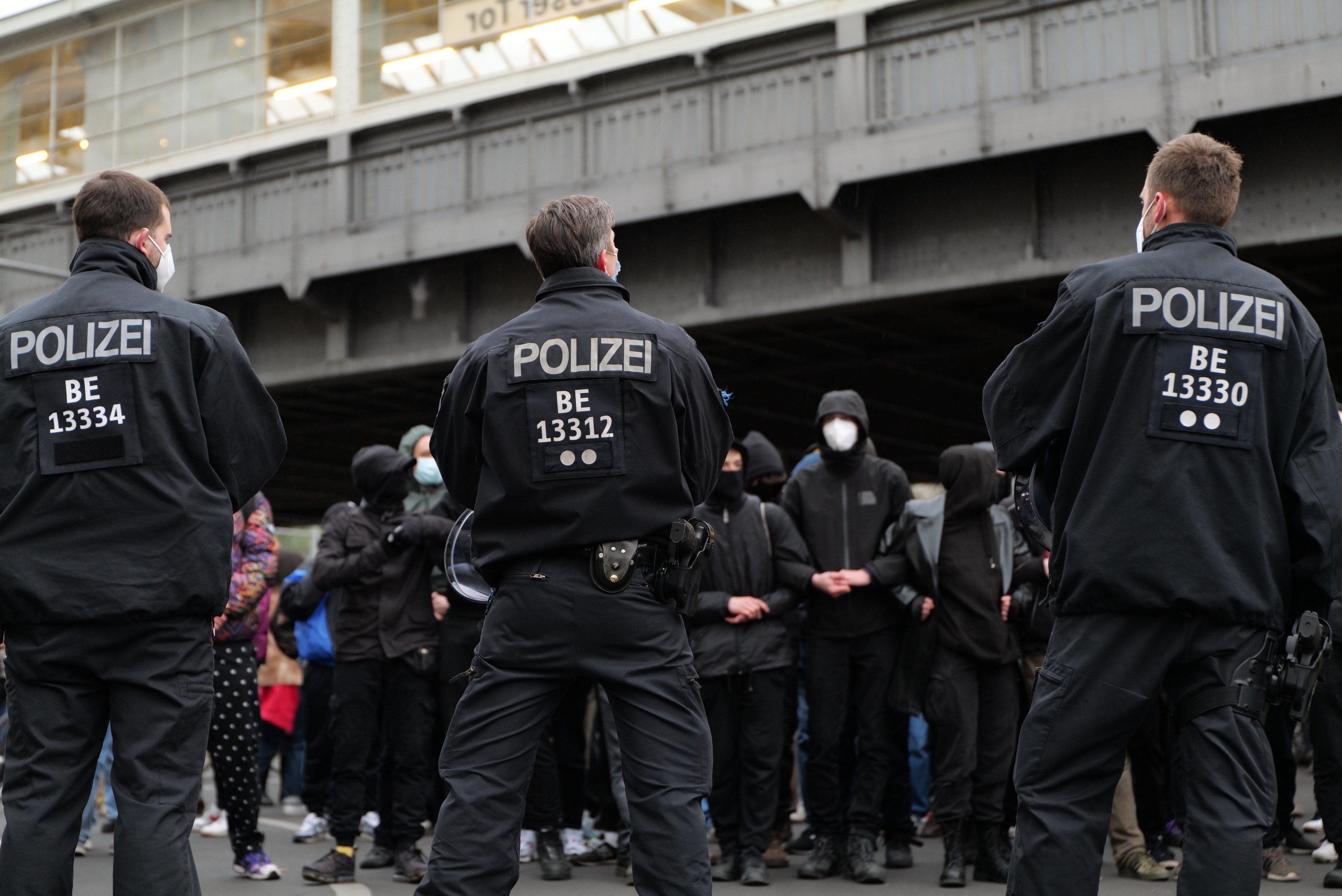 Polizeibeamte in schwarzen Uniformen und Masken stehen vor einer Menge, mit einer Brücke und einem Gebäude im Hintergrund während einer Demonstration gegen deutsche Anti-Terror-Maßnahmen.