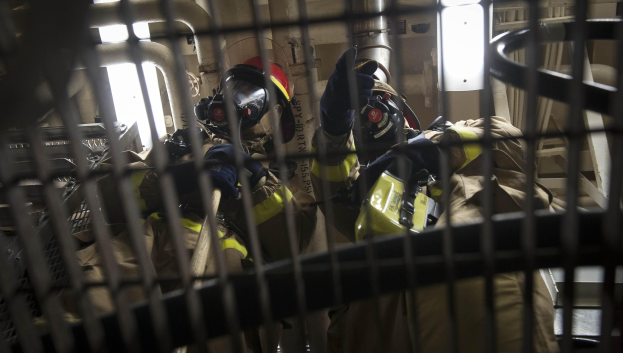 Eine Gruppe von Feuerwehrleuten in Schutzausrüstung steht in einem Raum mit einem Metallgitter im Vordergrund und verschiedenen Rohren und Stangen im Hintergrund, was darauf hinweist, dass sie auf einen Brand reagieren.