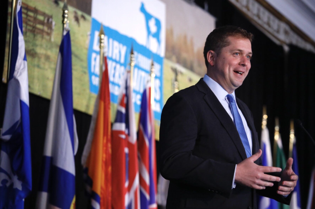 Ein Mann in Anzug und Krawatte steht an einem Rednerpult mit Mikrofon und spricht vor Flaggen und einem Bildschirm, wahrscheinlich spricht er über eine Regierungszahlung an die Milchindustrie.