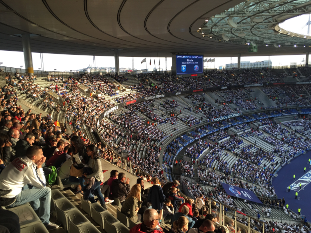 Großes Publikum in einem Stadion bei einem Fötballspiel mit einer Bühne auf der rechten Seite, Fahnen, Masten, einem Bildschirm und der Allianz Arena in München, Deutschland im Hintergrund.
