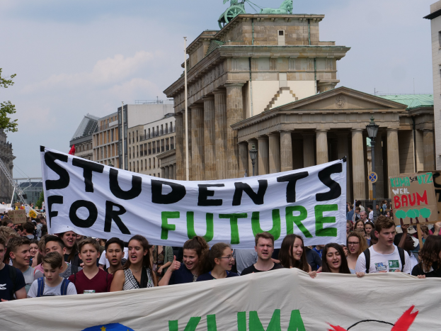 Gruppe von Schülern marschiert in Berlin mit einem leuchtend bunten 'Students for Future' Banner vor Gebäuden, Bäumen und Himmel.