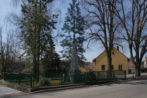 Eine Straße mit einem Holocaust-Denkmal in ihrer Mitte, umgeben von einem Metallzaun, umgeben von Gras, Bäumen, Häusern, einem Straßenmast und einem bewölkten Himmel.